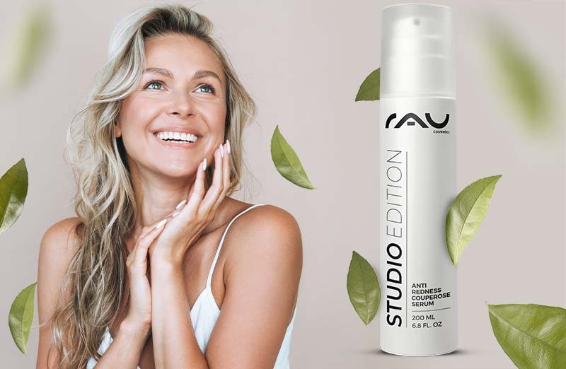 Lächelnde Frau mit langem, welligem Haar hält Anti-Rötung Serum von RAU Cosmetics vor grünen Blättern.