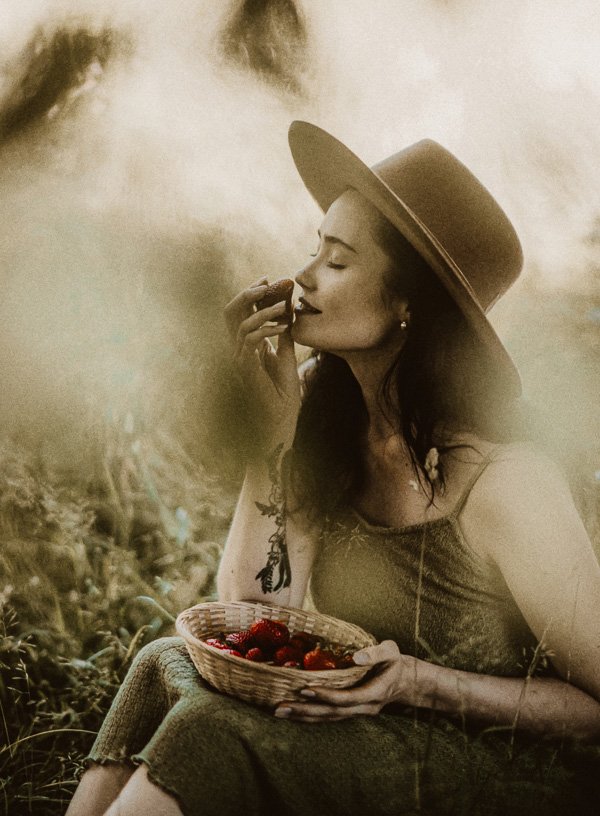 Frau mit Hut, die genussvoll eine Erdbeere aus einem Korb isst, symbolisiert somatische Intelligenz und bewusste Ernährung im Kontext von Gesundheit und Wohlbefinden.