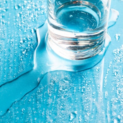 booster_trocken e_haut Glas mit Wasser auf blauer Oberfläche, umgeben von Wassertropfen, symbolisiert Feuchtigkeit und Pflege für trockene Haut, passend zum Thema Slugging in der Hautpflege.