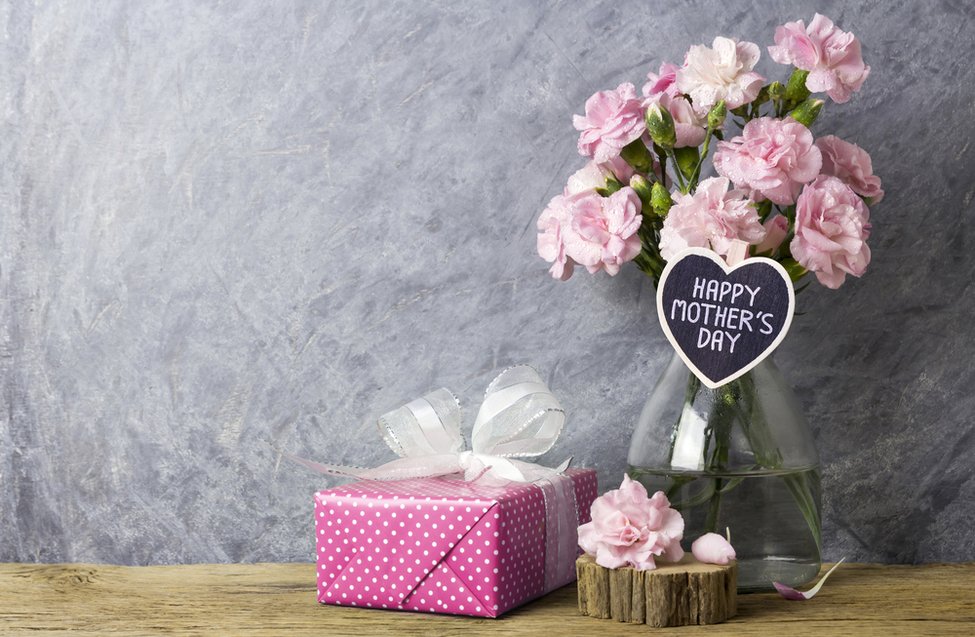 Blumenstrauß mit rosa Nelken in einer Glasvase, beschriftet mit "HAPPY MOTHER'S DAY", neben einem pinken Geschenk mit weißen Punkten auf einem Holztisch, passend zum Muttertag und zu RAU Cosmetics' Angeboten für Pflegeprodukte.
