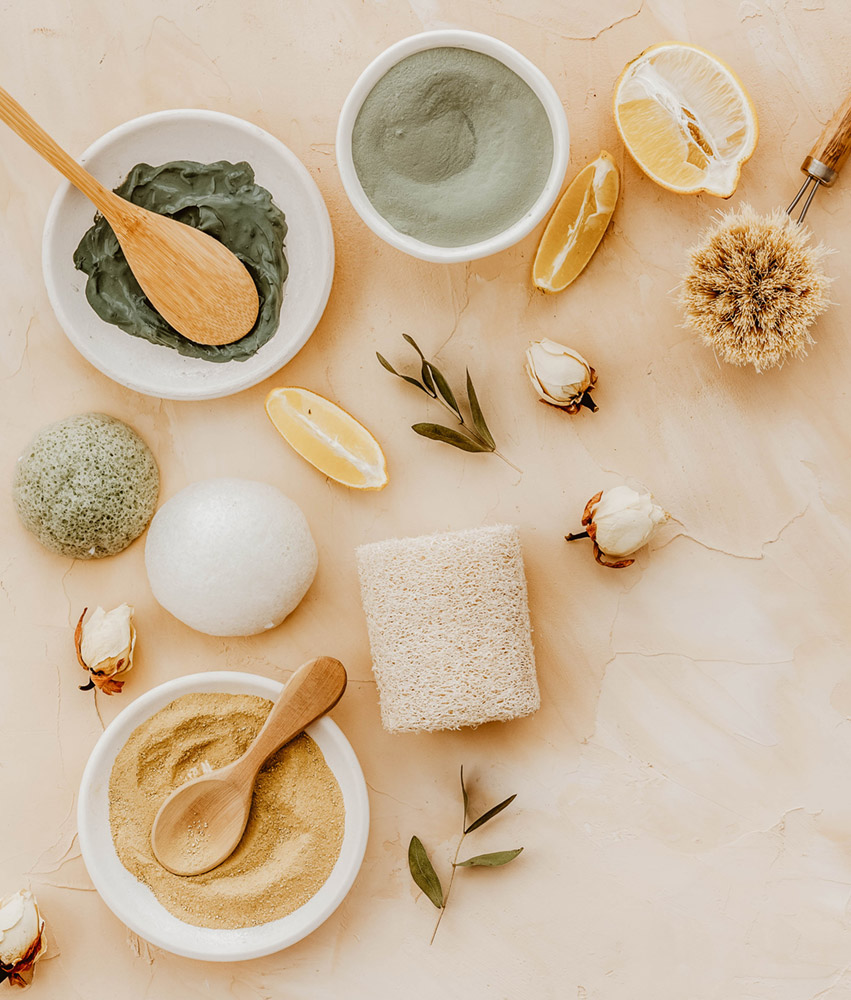 vegane-kosmetik-grunde_rau-cosmetics Vegane Kosmetikprodukte, darunter grüne und graue Masken, Peeling-Schwämme, Zitrusfrüchte und natürliche Inhaltsstoffe, auf einem hellen Untergrund, umgeben von Pflanzen und Rosenblüten.