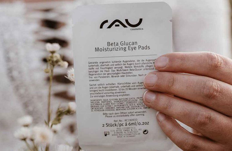 Die Verpackung der RAU Cosmetics Beta Glucan Moisturizing Eye Pads wird von einer Person gehalten und hebt die hautpflegenden Vorteile von Beta Glucan hinsichtlich der Feuchtigkeitsversorgung und des Hautschutzes hervor.