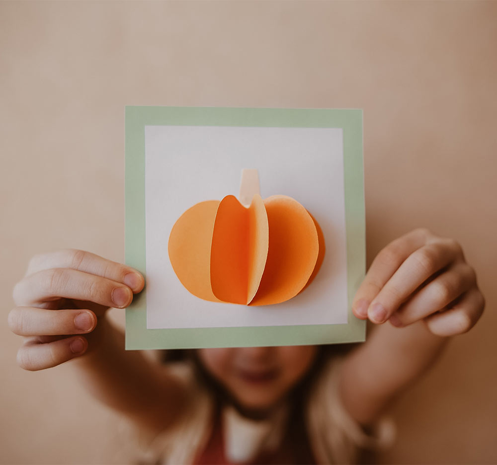 regen-beschaeftigung-kinder-familie-basteln-spass Kind hält ein handgemachtes Kürbis-Kunstwerk aus Papier und zeigt damit herbstliche Kreativität für Indoor-Aktivitäten an regnerischen Tagen.
