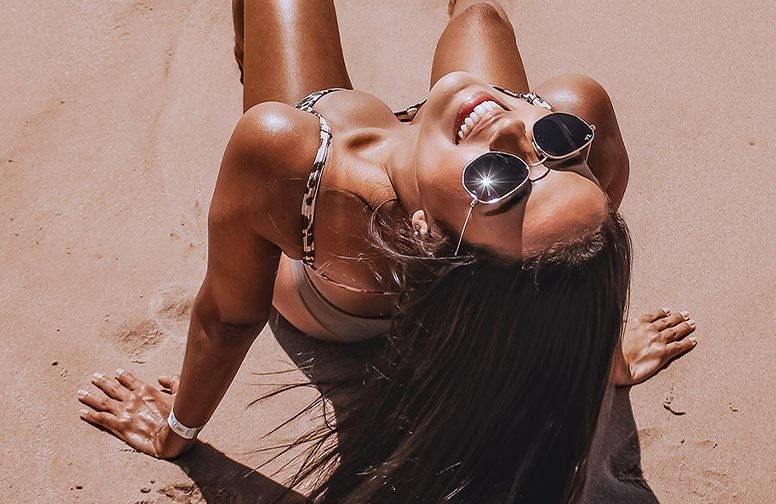 Frau mit Sonnenbrille und sonnengebräunter Haut, lächelnd am Strand, symbolisiert Sommerurlaub und Hautpflege für langanhaltende Bräune.