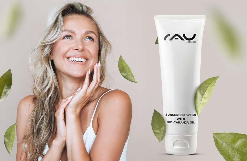 Frau mit freudigem Gesicht und Sonnenschutzmittel SPF 50 mit Bio-Karanja-Öl von RAU Cosmetics vor grünen Blättern.