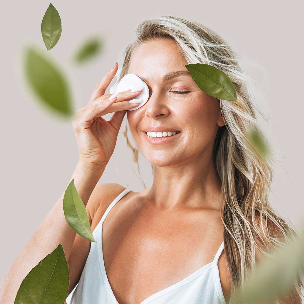 Frau mit blonden Haaren, die sanft ein Wattepad auf ihr Gesicht aufträgt, um Hautpflege zu demonstrieren, umgeben von grünen Blättern, symbolisiert natürliche Inhaltsstoffe und Hautgesundheit.