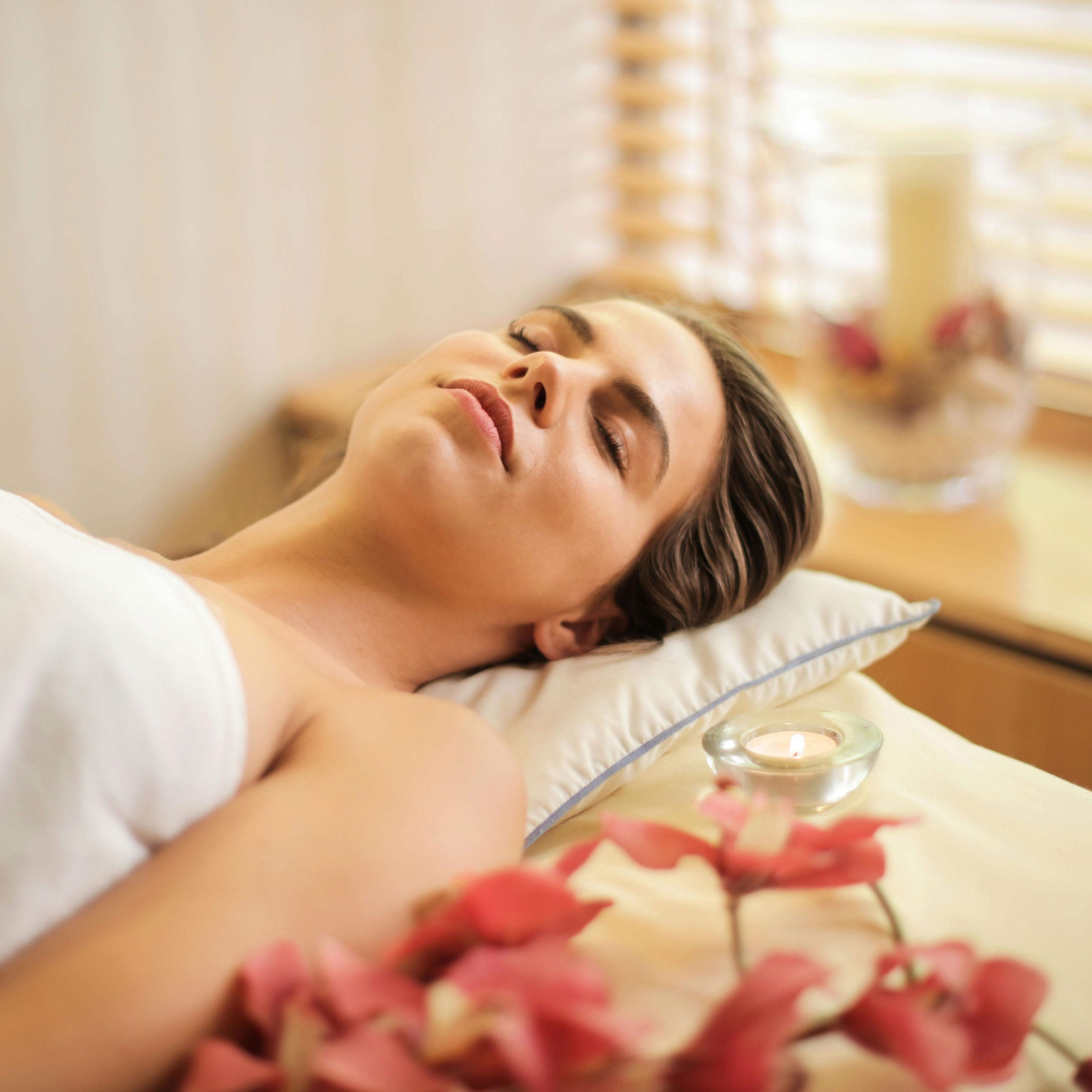 pexels-olly-3757942 Frau entspannt während einer Gesichtsbehandlung mit Blumen und Kerze, repräsentiert die Vorteile der Buccal Massage für Hautverjüngung und Entspannung.