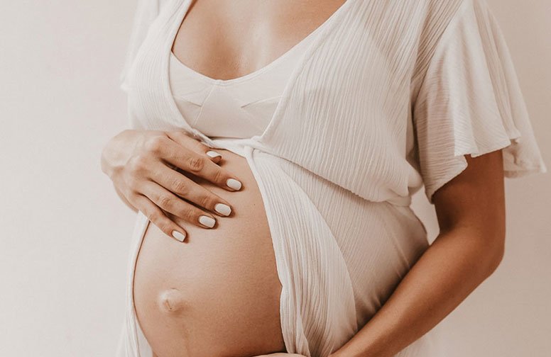 Schwangere Frau in weißem Kleid, die ihren Bauch sanft hält, symbolisiert Hautpflege und Veränderungen während der Schwangerschaft.