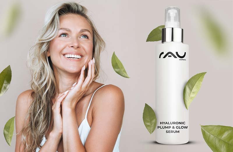 Frau mit blonden Wellenhaaren und Hyaluronsäure Plump & Glow Serum von RAU, umgeben von grünen Blättern.